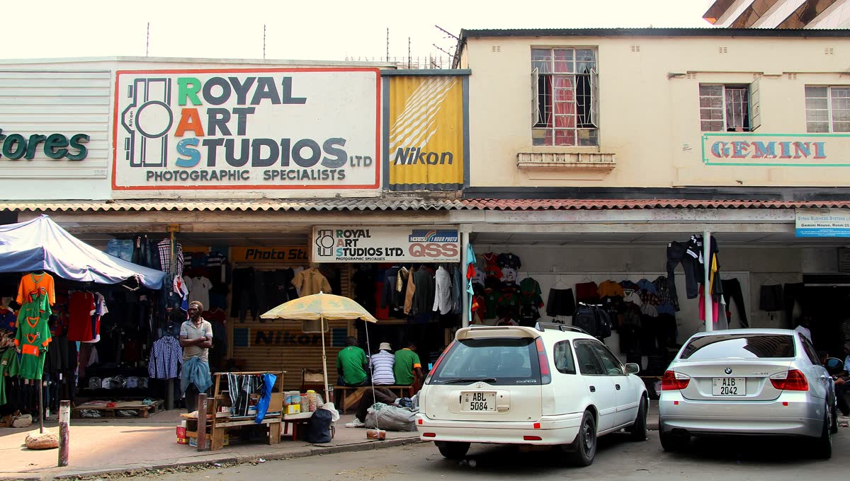 Shopfronts on Cairo Road, Lusaka, with signs above, clothes on display and cars parked in front